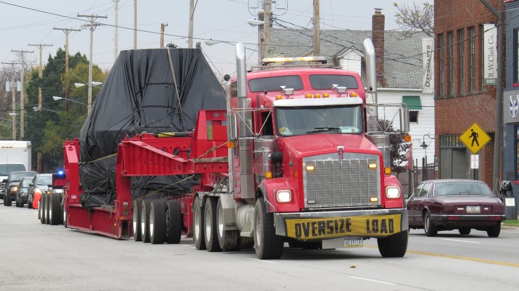 Red Kenworth semi-truck pulling a multi-axle heavy-haul lowboy with a tarped oversize load